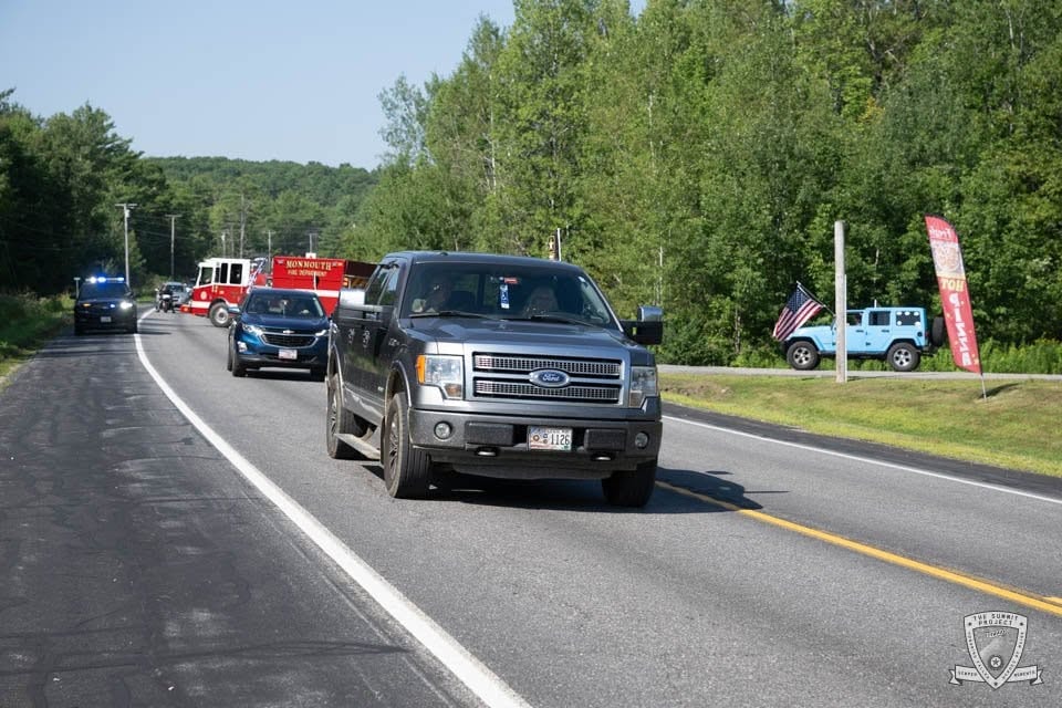 A gray Ford F-150 rolls through the procession with the blue Jeep and fire truck visible behind.